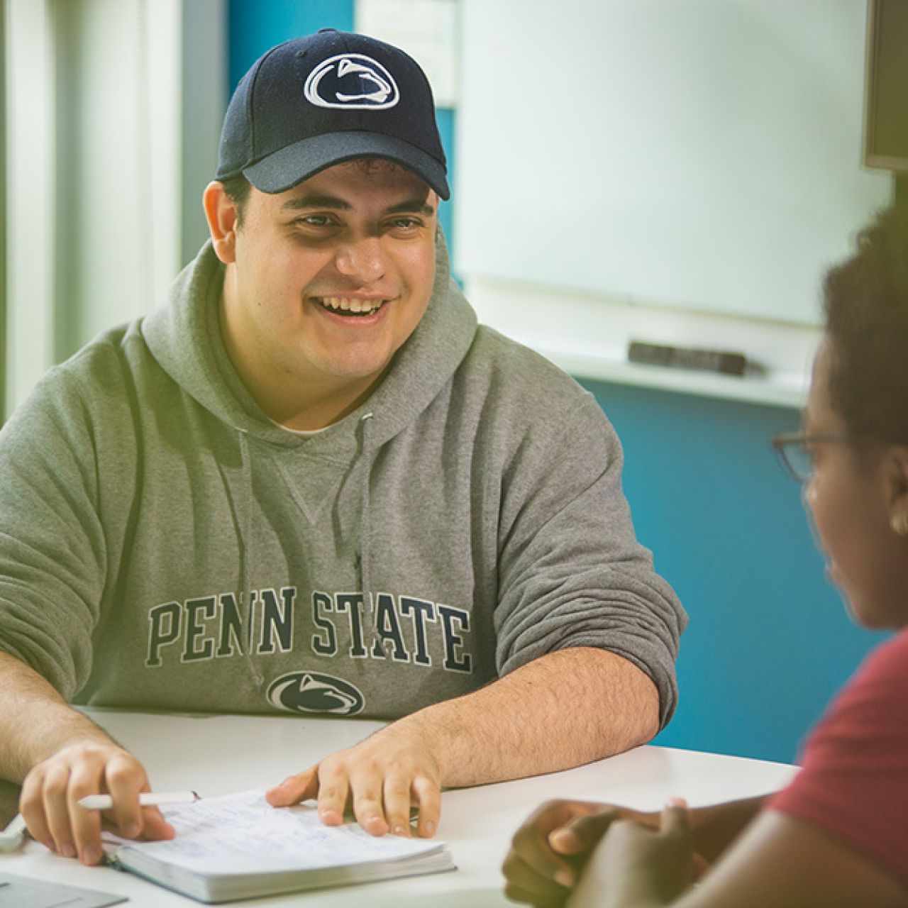 student in Penn State hoodie and cap talking to other student across table