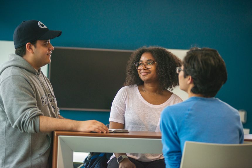 one student standing, two students seated at table, having conversation